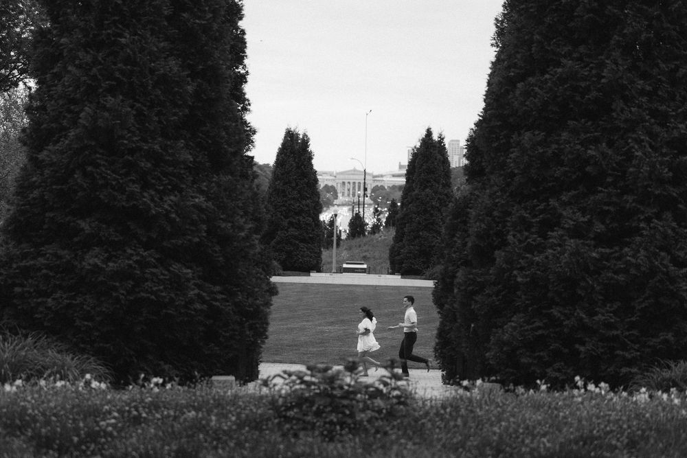 Black and white photo of a couple jogging through a tree-lined Chicago Maggie Daley park path with a building visible in the distance.