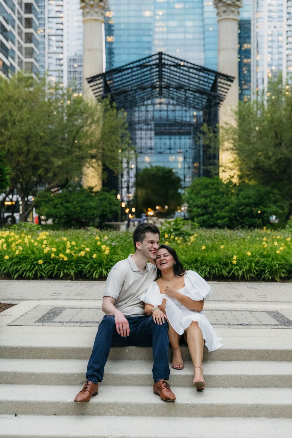 Couple sitting on steps in Maggie Daley Park Chicago, with lush gardens and modern glass building in background during engagement session