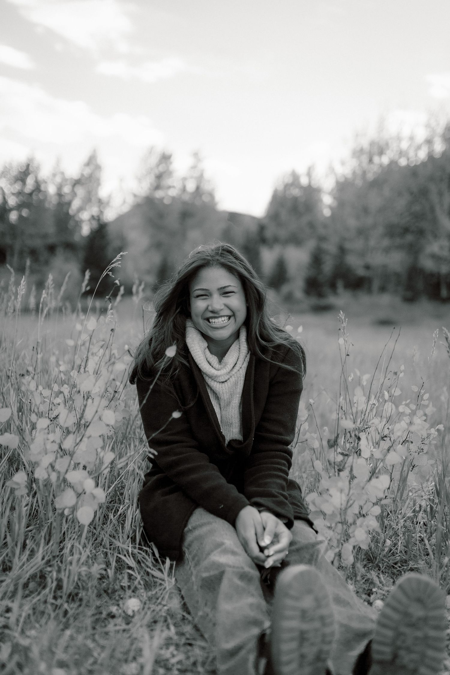 Black and white photo of a smiling young woman sitting in a meadow field with trees and mountains in the background.
