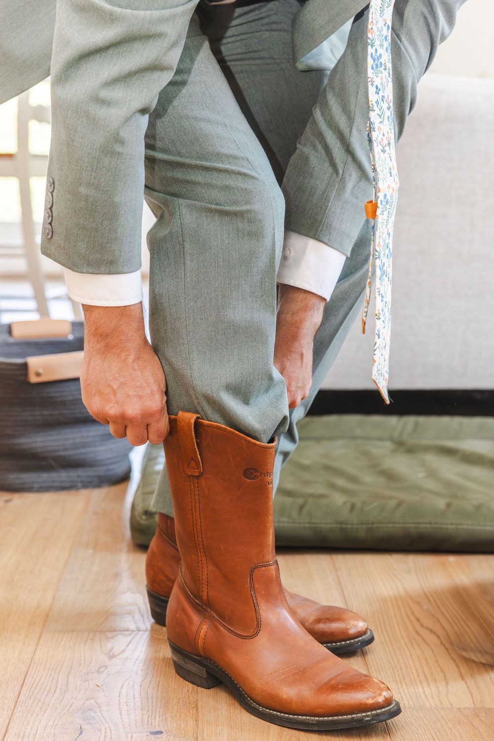 Groom in gray suit pulling on brown leather cowboy boots while getting dressed for his wedding day.