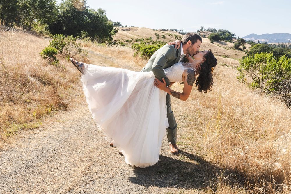 Groom dips bride in white gown for a romantic kiss on a sunny dirt path surrounded by golden hills.