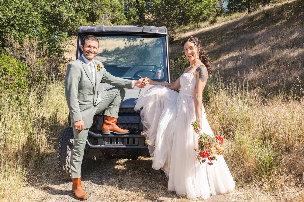 Bride and groom posing together in front of a black off-road vehicle on a sunny wooded trail.