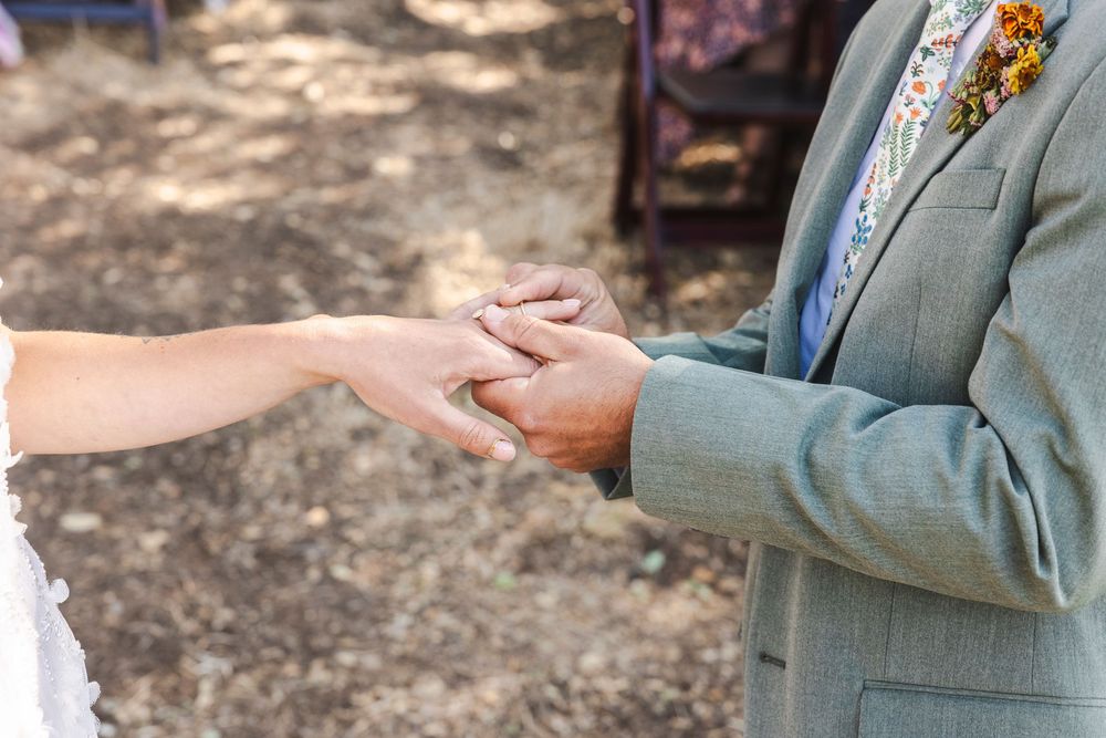 Groom in gray suit holds bride's hand during outdoor wedding ring exchange ceremony.