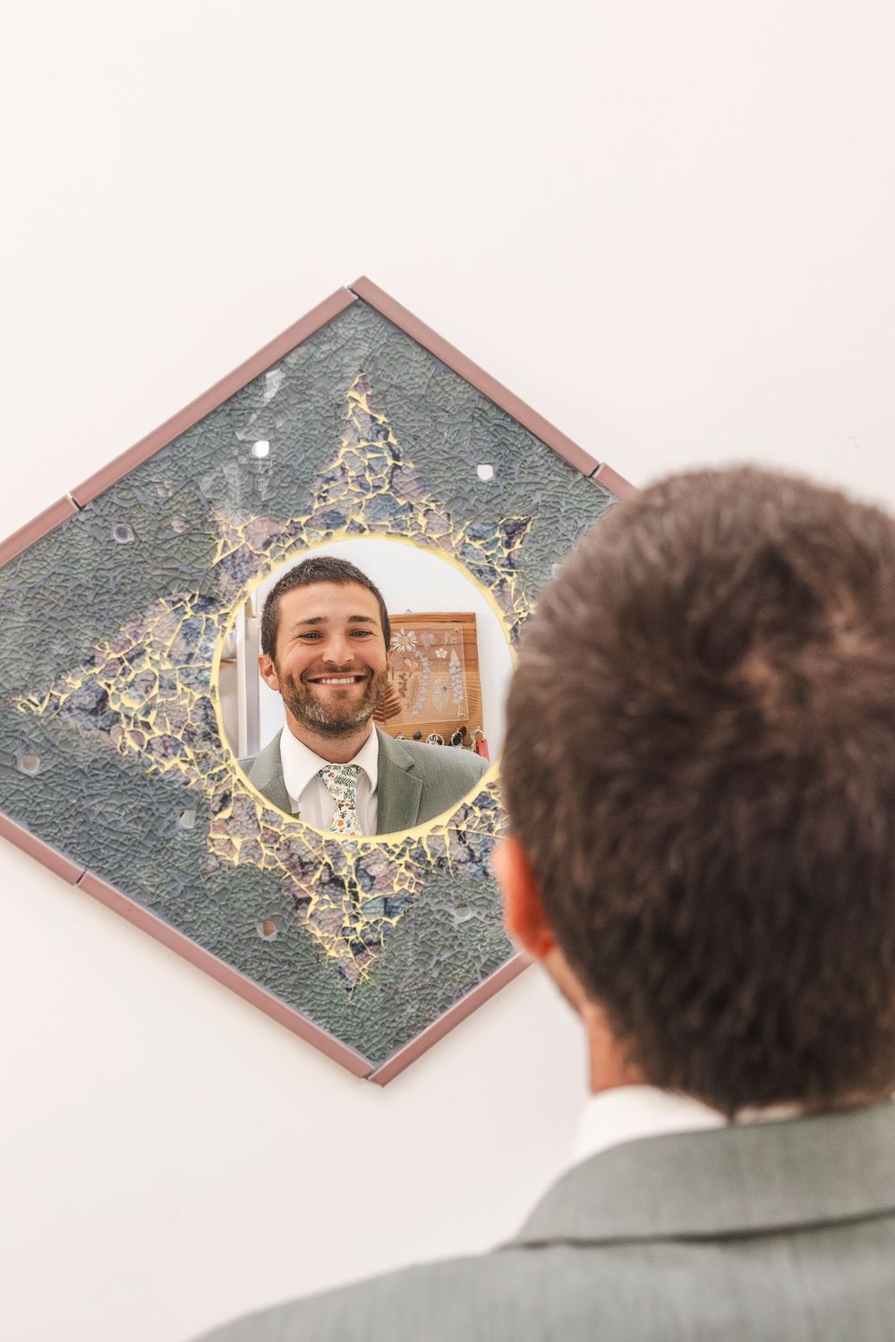 A smiling man in a gray suit looks at his reflection in a decorative round mirror mounted on a white wall.
