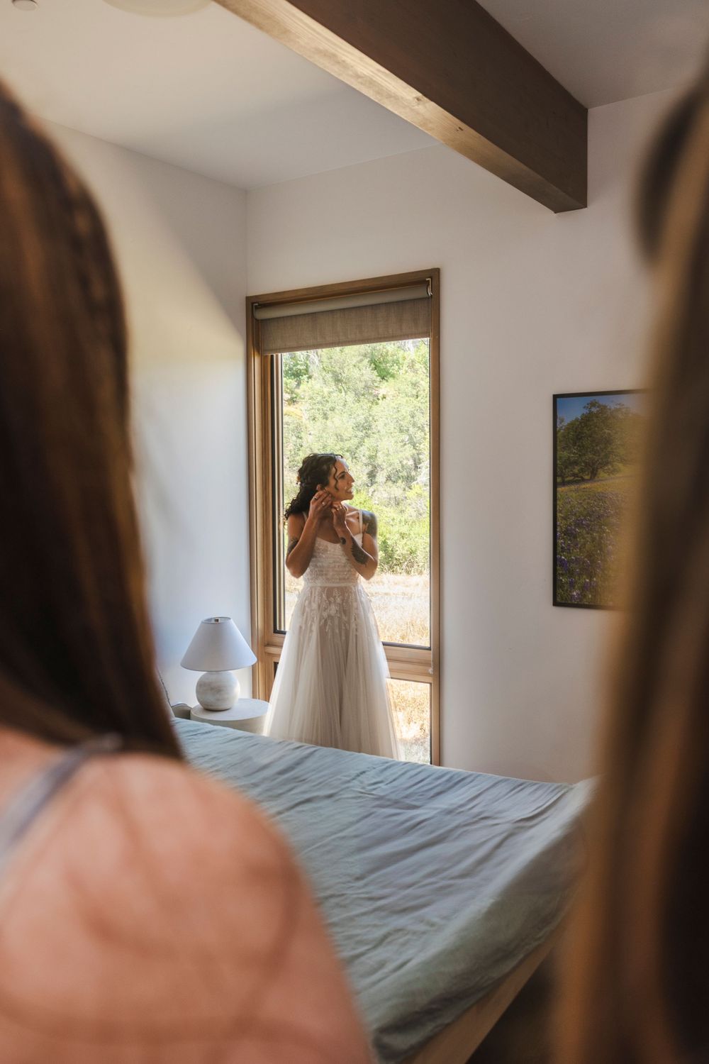 Bride in white wedding dress standing by a sunlit window, holding a bouquet, viewed from behind guests in a cozy bedroom.