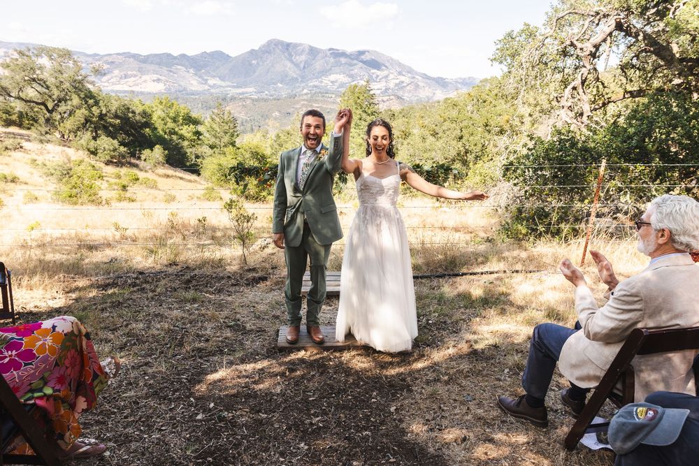 Joyful newlyweds raise hands at outdoor mountain wedding ceremony with guests applauding in scenic California hillside setting.