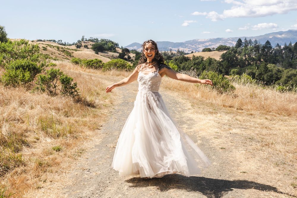 Bride in a flowing white wedding gown twirling on a dirt path with golden hills and mountains in the background.