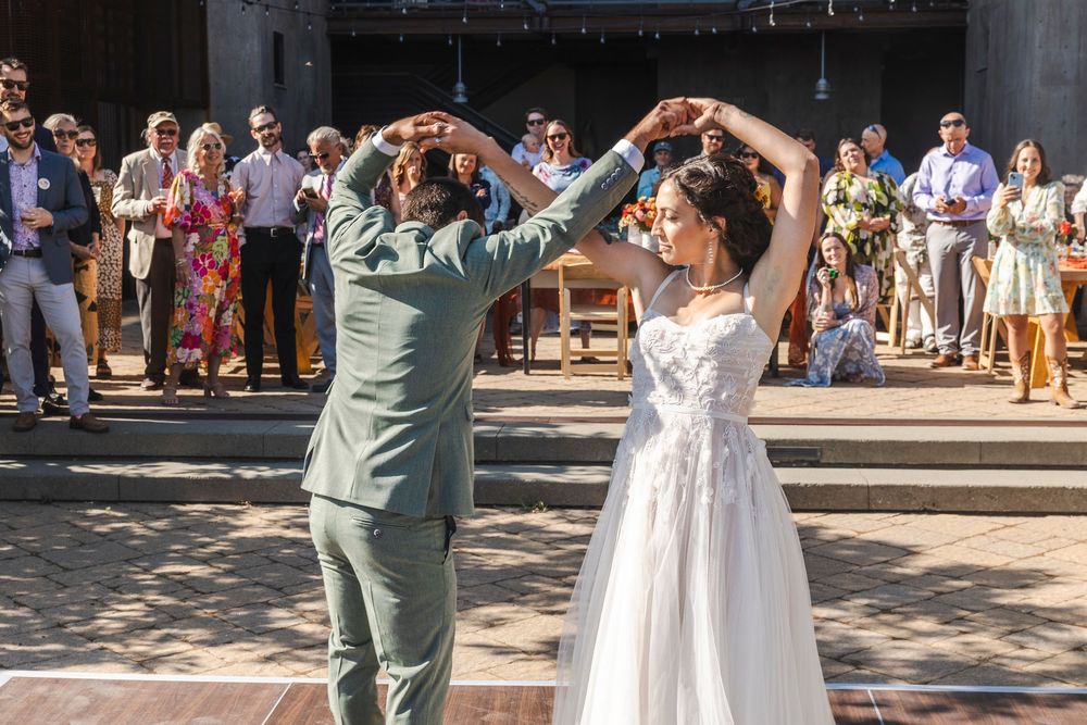 Bride and groom share first dance outdoors at sunny wedding reception with guests applauding in background.