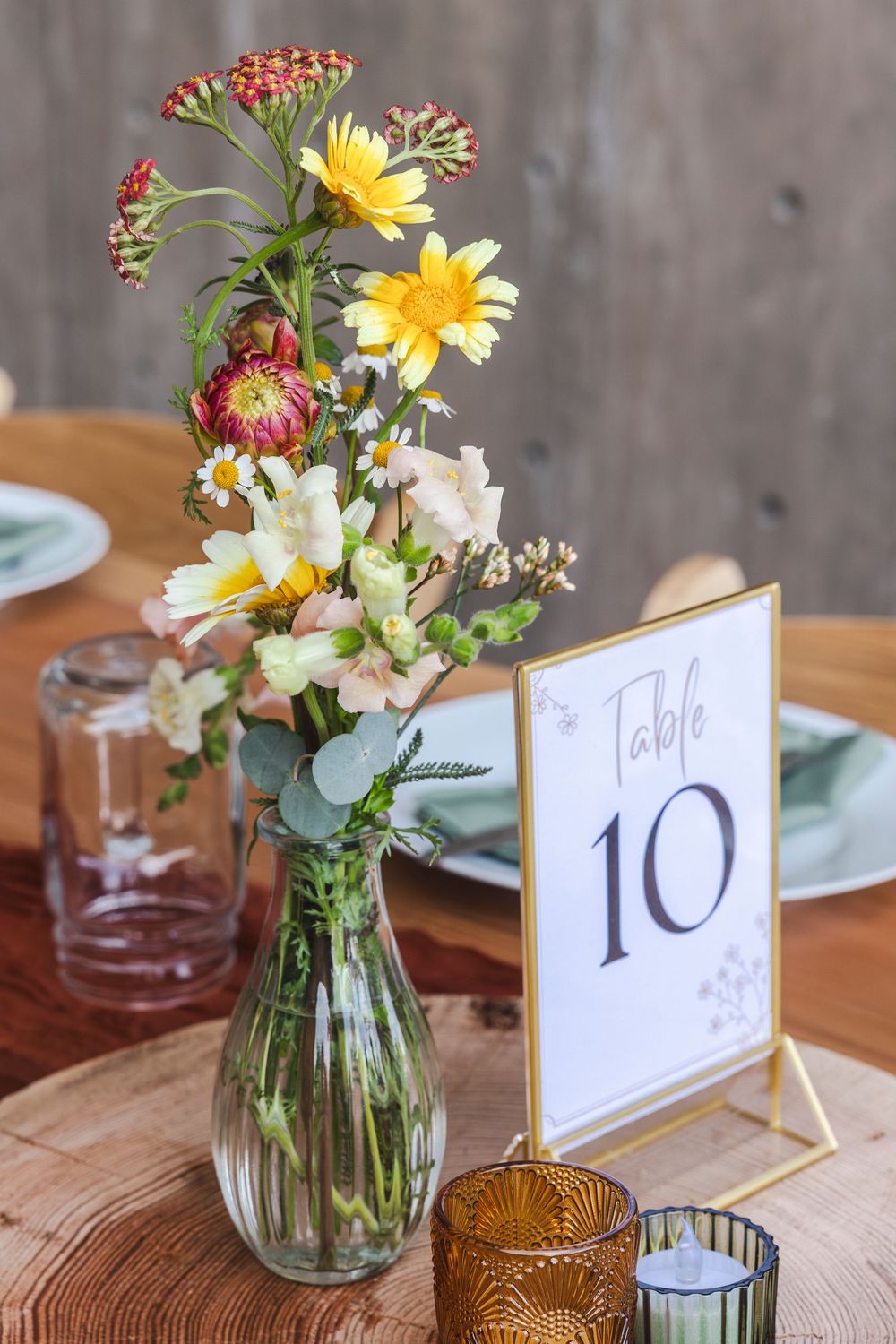 Rustic wedding table centerpiece with wildflowers in glass vase beside Table 10 sign on wooden slice.