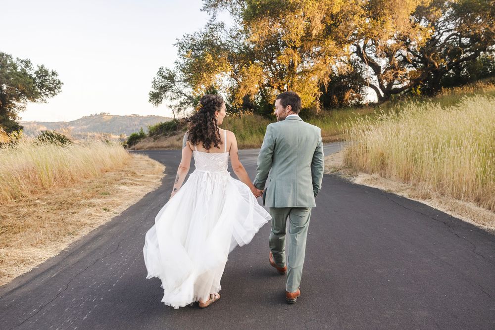 Bride and groom walking hand in hand down a scenic road surrounded by golden hills and trees.