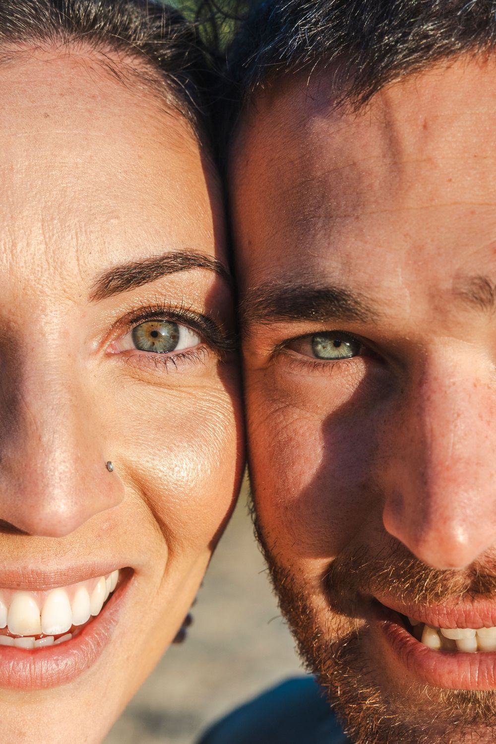 Close-up portrait of a smiling couple with green eyes, cheeks touching, looking at the camera in natural sunlight.