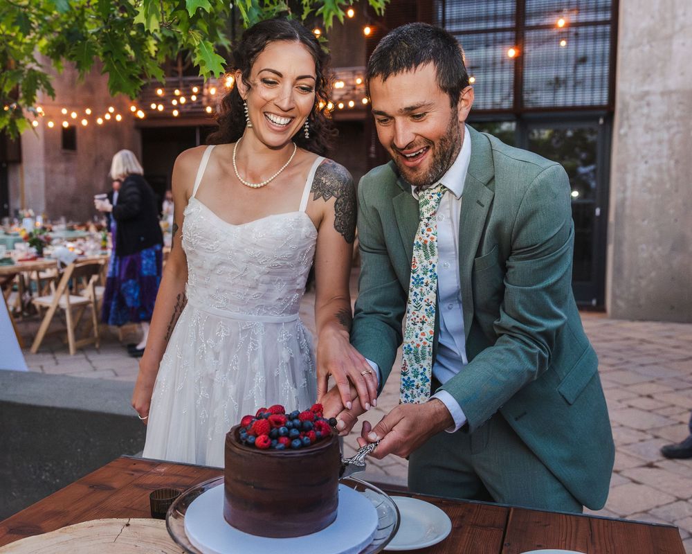 Smiling bride and groom in a green suit cutting a chocolate cake topped with fresh berries at their wedding.