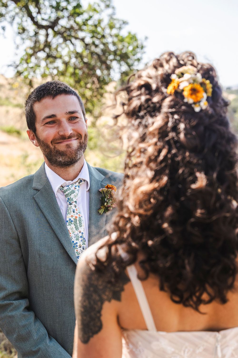 Smiling groom in gray suit with floral tie and boutonniere faces bride with curly hair and yellow flowers outdoors.