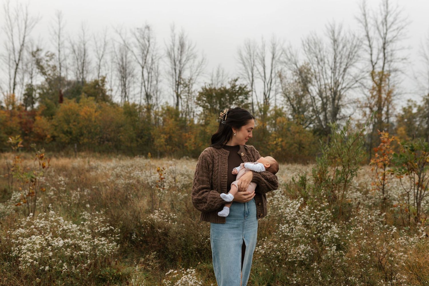 Mother holding newborn baby in autumn field with colorful fall foliage background.
