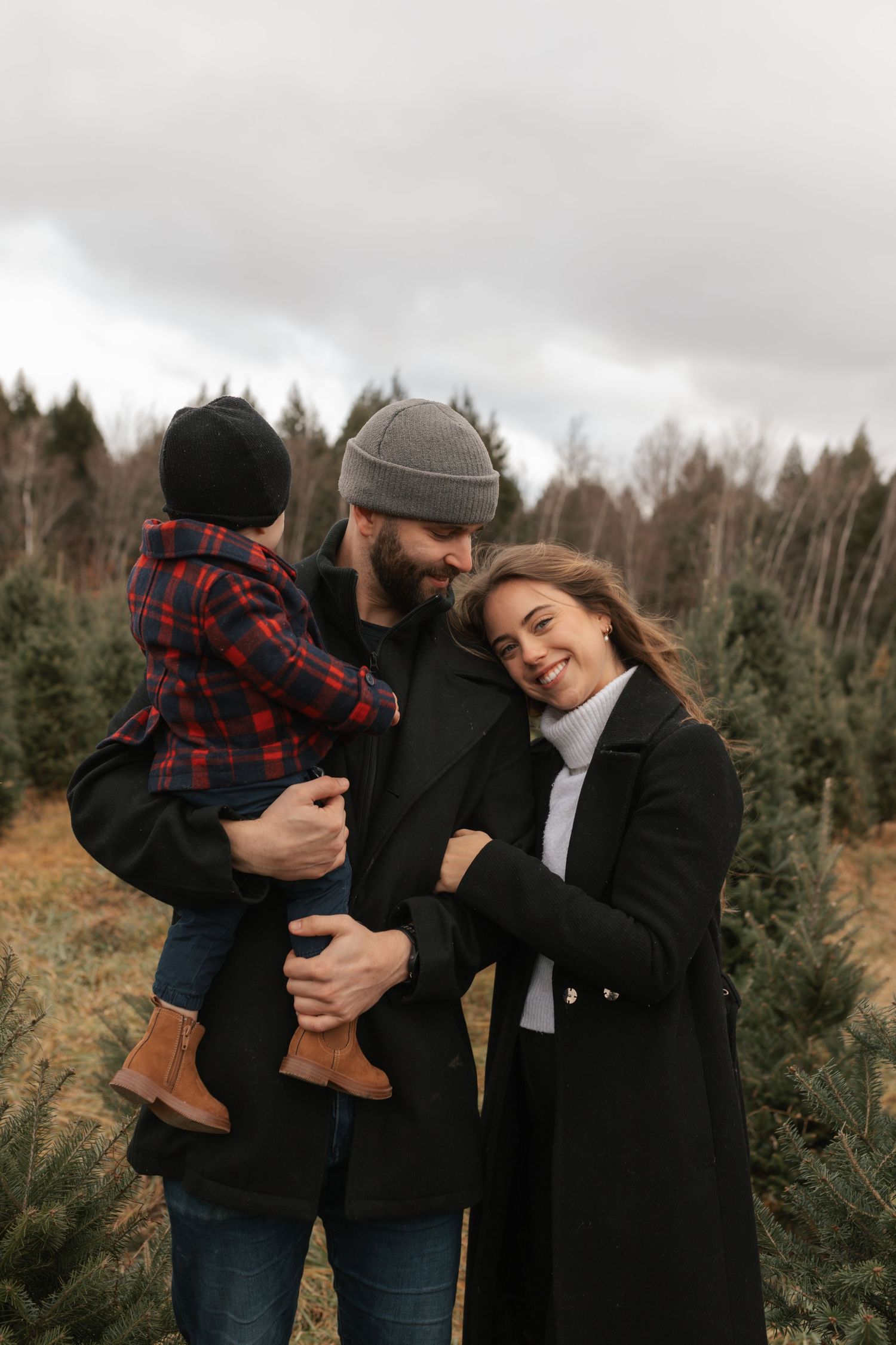 Happy family with bearded father holding child and smiling woman in black coat at Christmas tree farm.