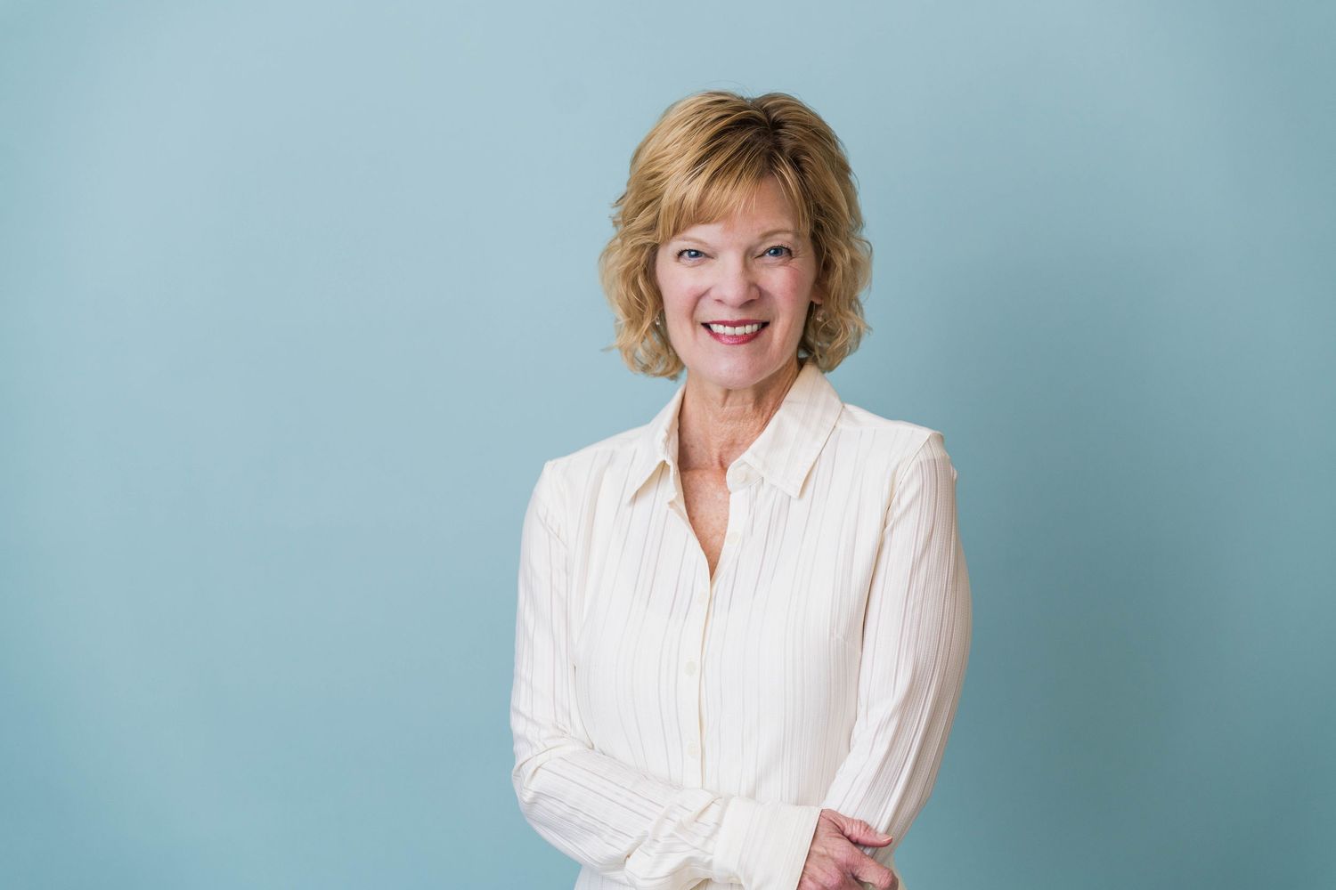 Smiling middle-aged woman with short blonde hair wearing a white blouse, posing against a light blue background.