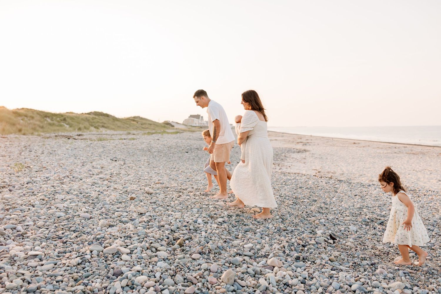 Family in white outfits walking on a rocky beach at sunset with two young children.