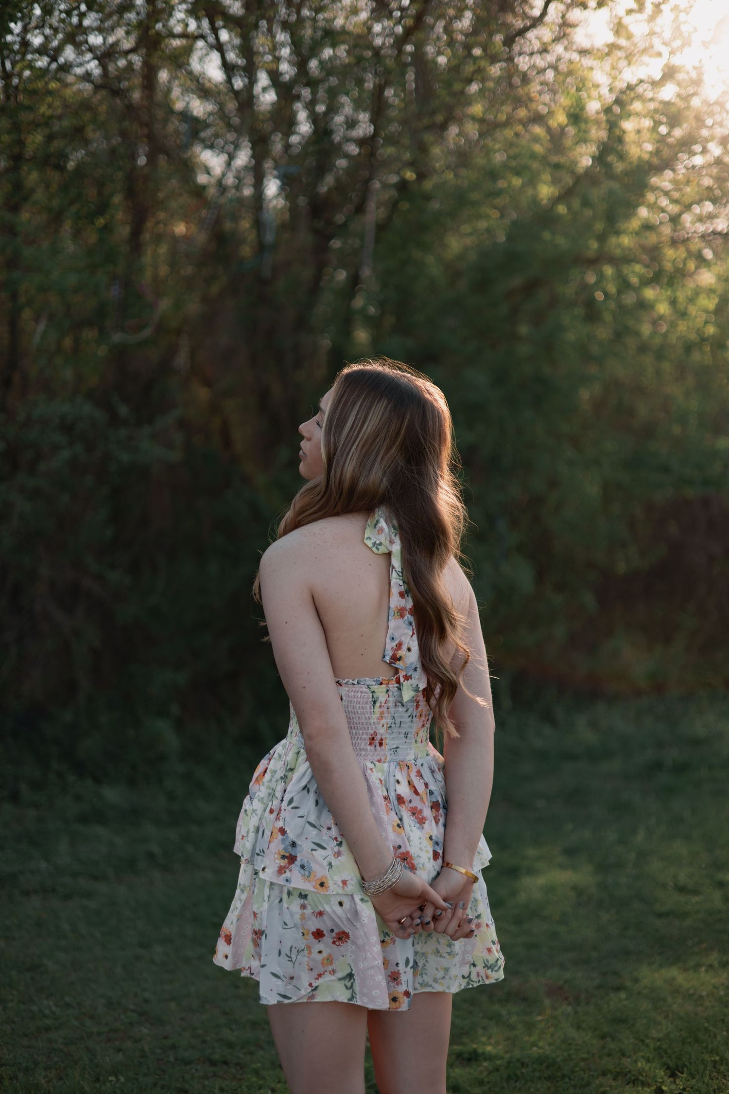 Young woman in floral sundress stands in golden hour sunlight with lush green trees in background.