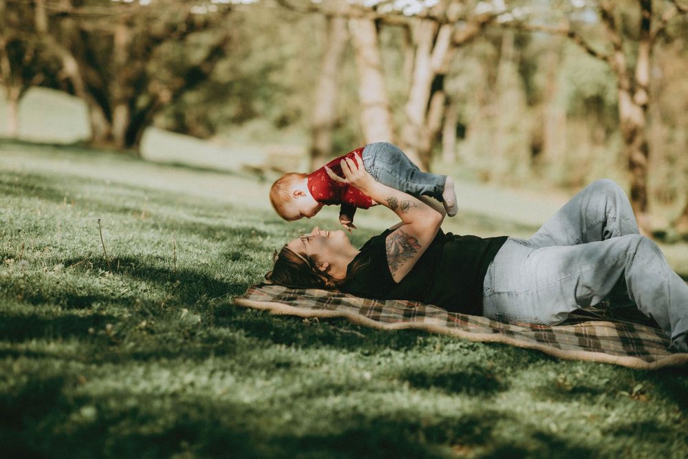Parent lying on blanket in park, playfully lifting smiling baby in red outfit outdoors on sunny day.