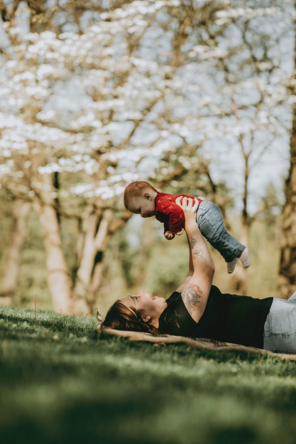 Mother lying on grass lifting baby in red outfit overhead near blooming spring trees.