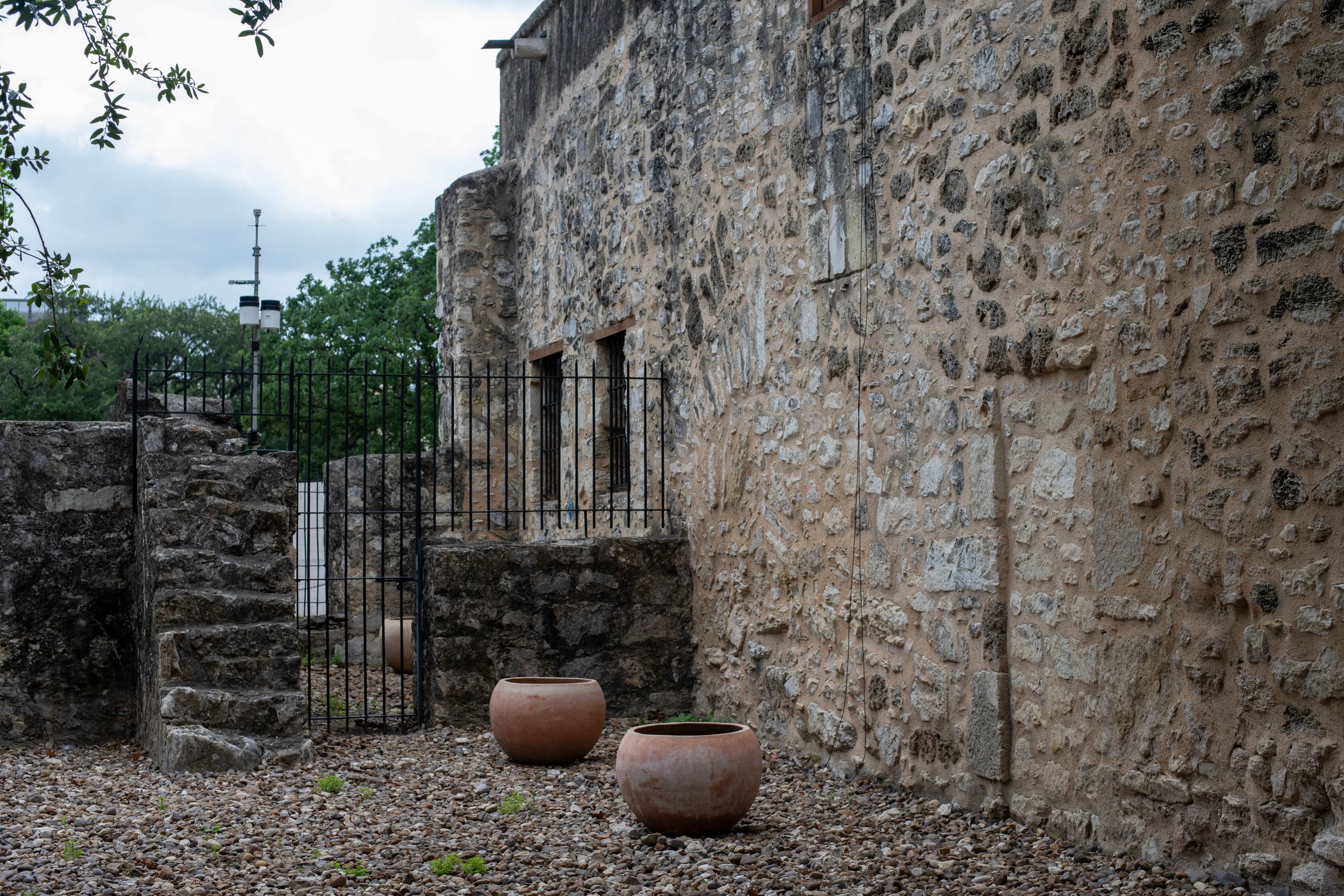 Historic stone mission wall with iron gate and two terracotta pots on gravel ground.