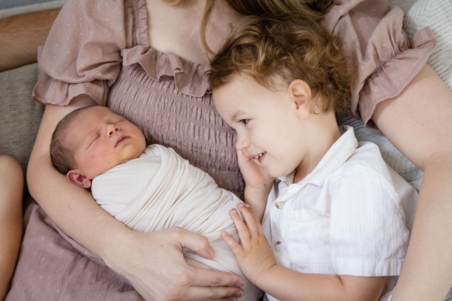 A mother holds her swaddled newborn while a young toddler boy gazes lovingly at the baby sibling.