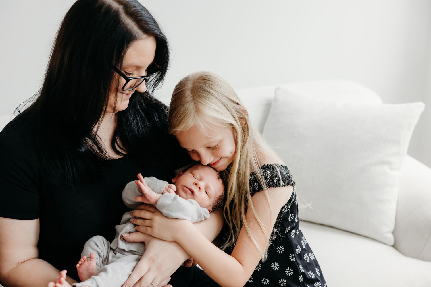 A mother with dark hair holds her newborn baby while a young blonde girl lovingly kisses the infant on a white sofa.