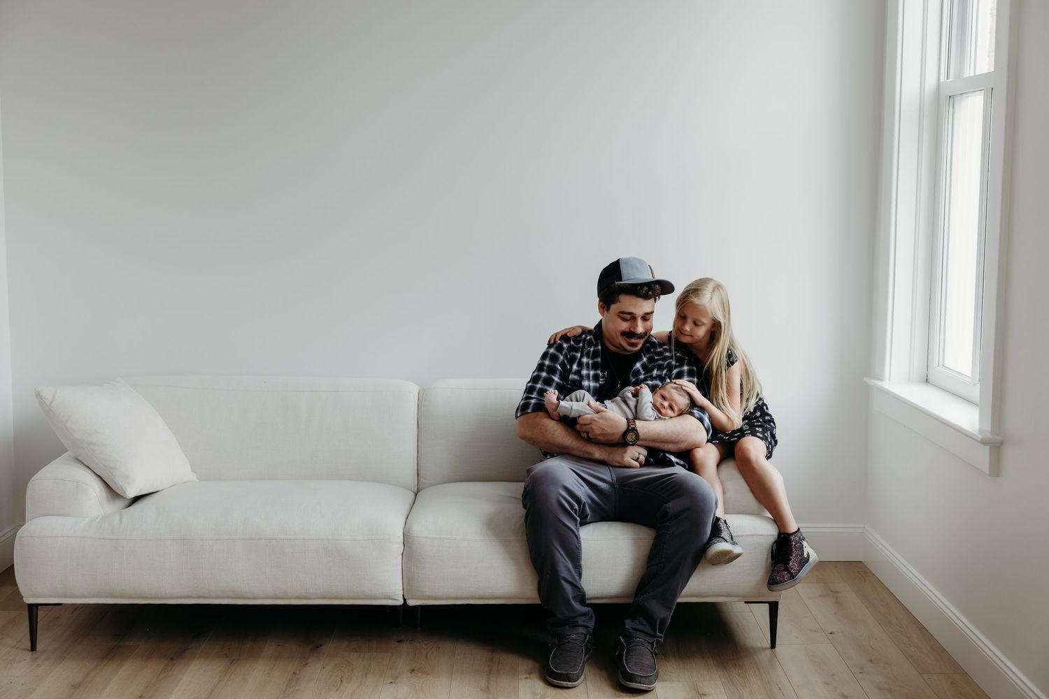 A happy family sits on a white sofa, with parents lovingly cradling their newborn baby in a bright, modern living room.