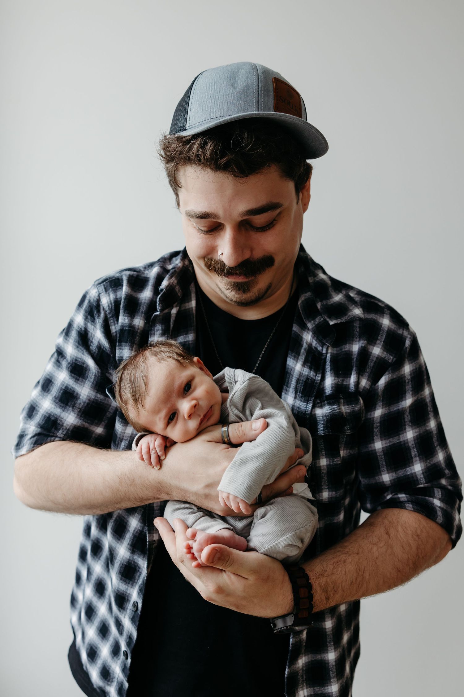 A young father with a mustache and cap tenderly holds his sleeping newborn baby close to his chest.