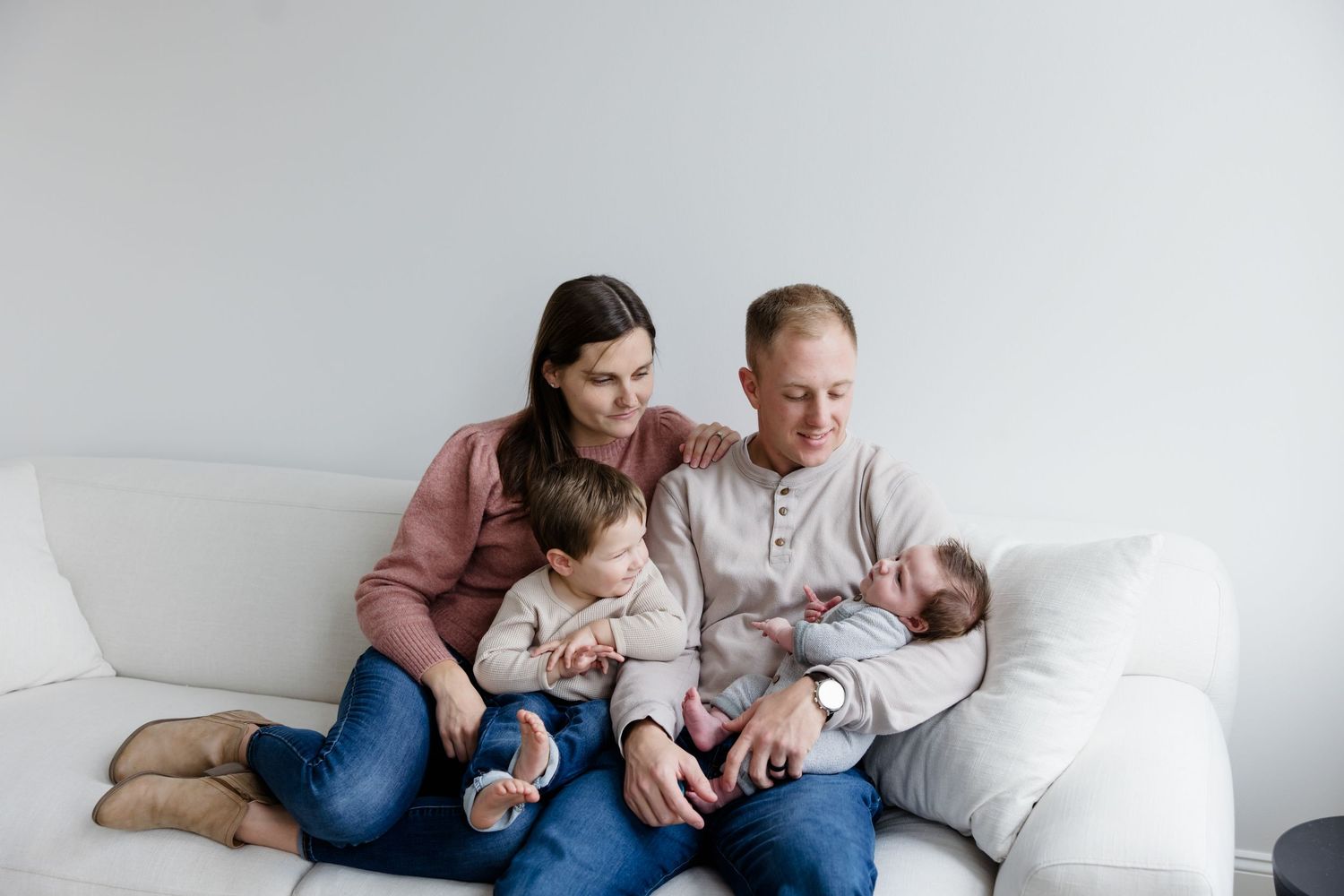 Family of four sitting on white couch, parents smiling at newborn baby while toddler sits between them.