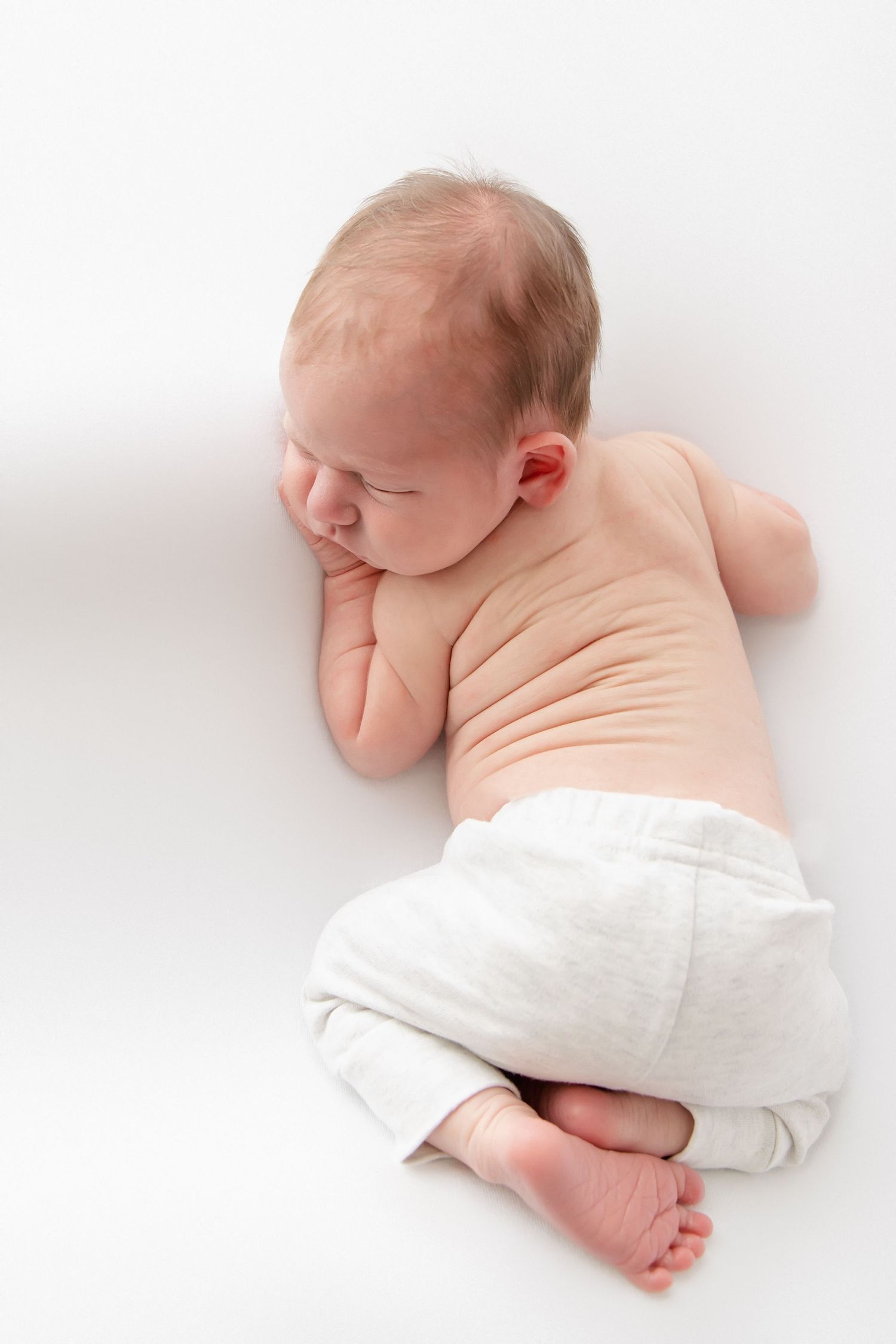 A peaceful newborn baby sleeping on its side wearing white pants on a soft white background.