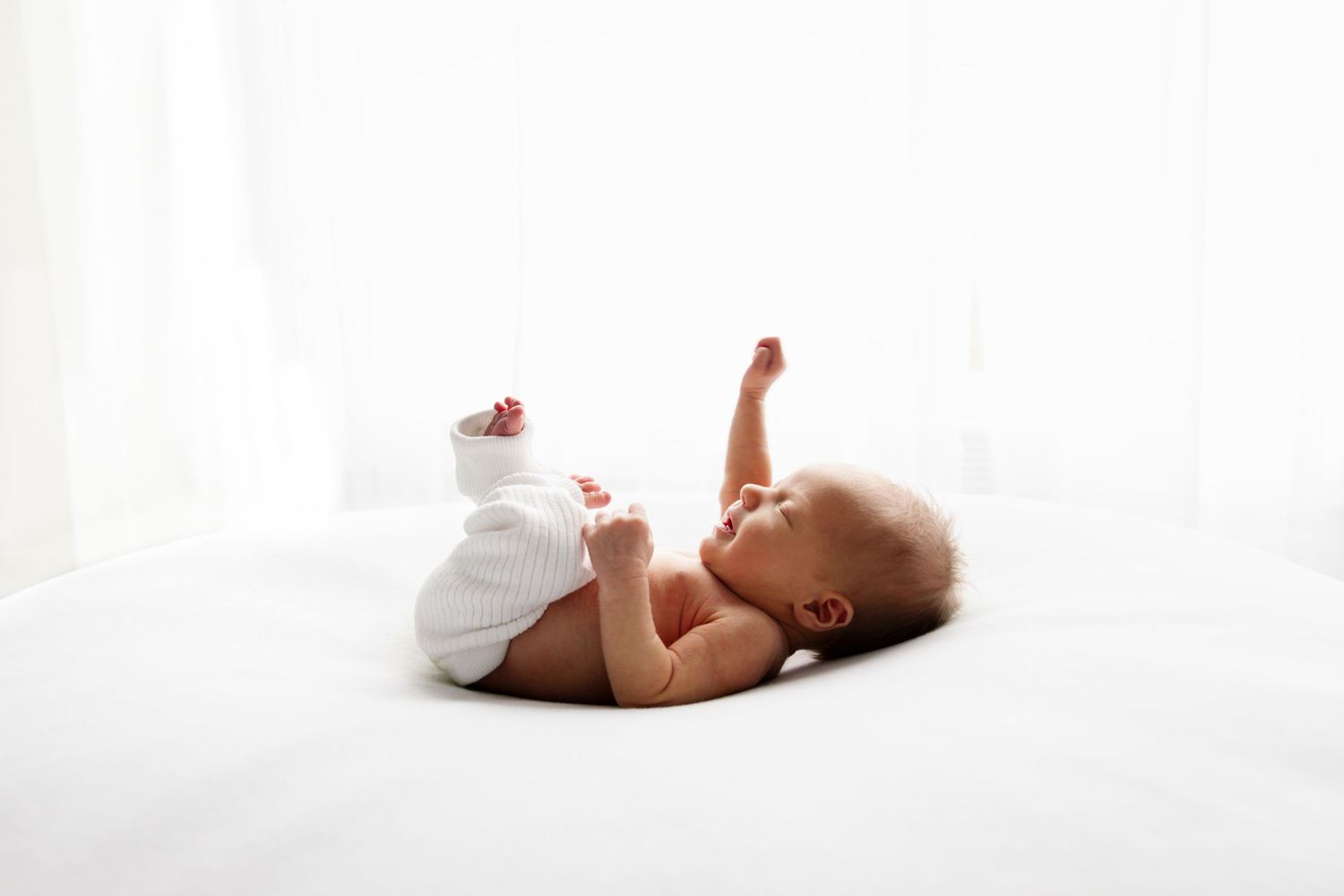 A peaceful newborn baby lying on a white bed in a diaper, stretching arms and legs in a bright, airy room.