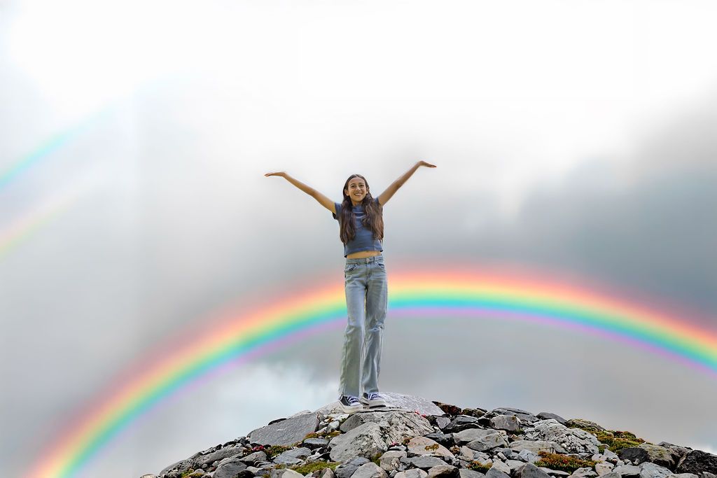 Young woman standing triumphantly on rocky mountain peak with arms raised, framed by a vibrant double rainbow in cloudy sky.