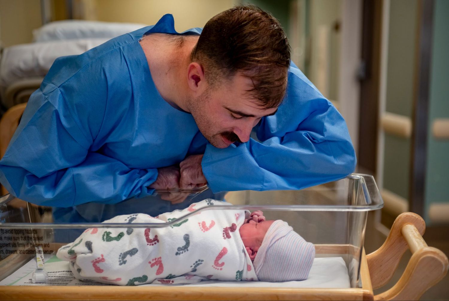 A parent in blue hospital attire leans over a newborn baby in a hospital bassinet displaying intense emotion.