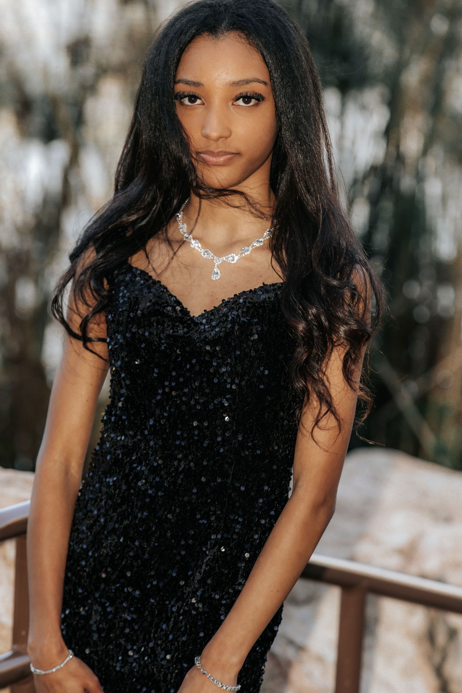 Young woman in black sequin dress with silver necklace and bracelet poses outdoors near wooden railing.