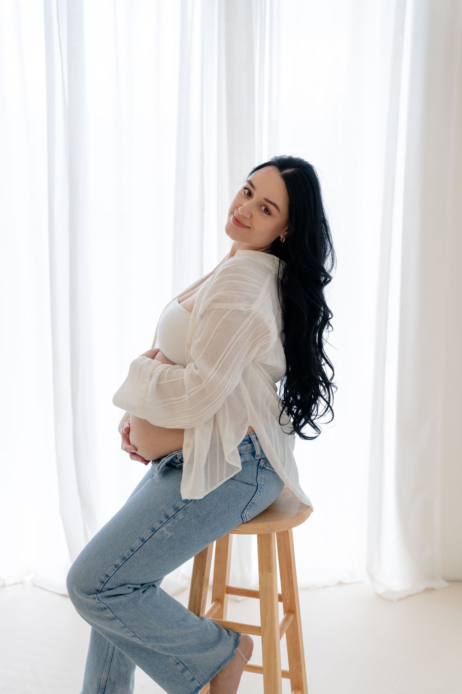 Pregnant woman with dark hair sits on wooden stool in white shirt and jeans, cradling baby bump in bright studio.