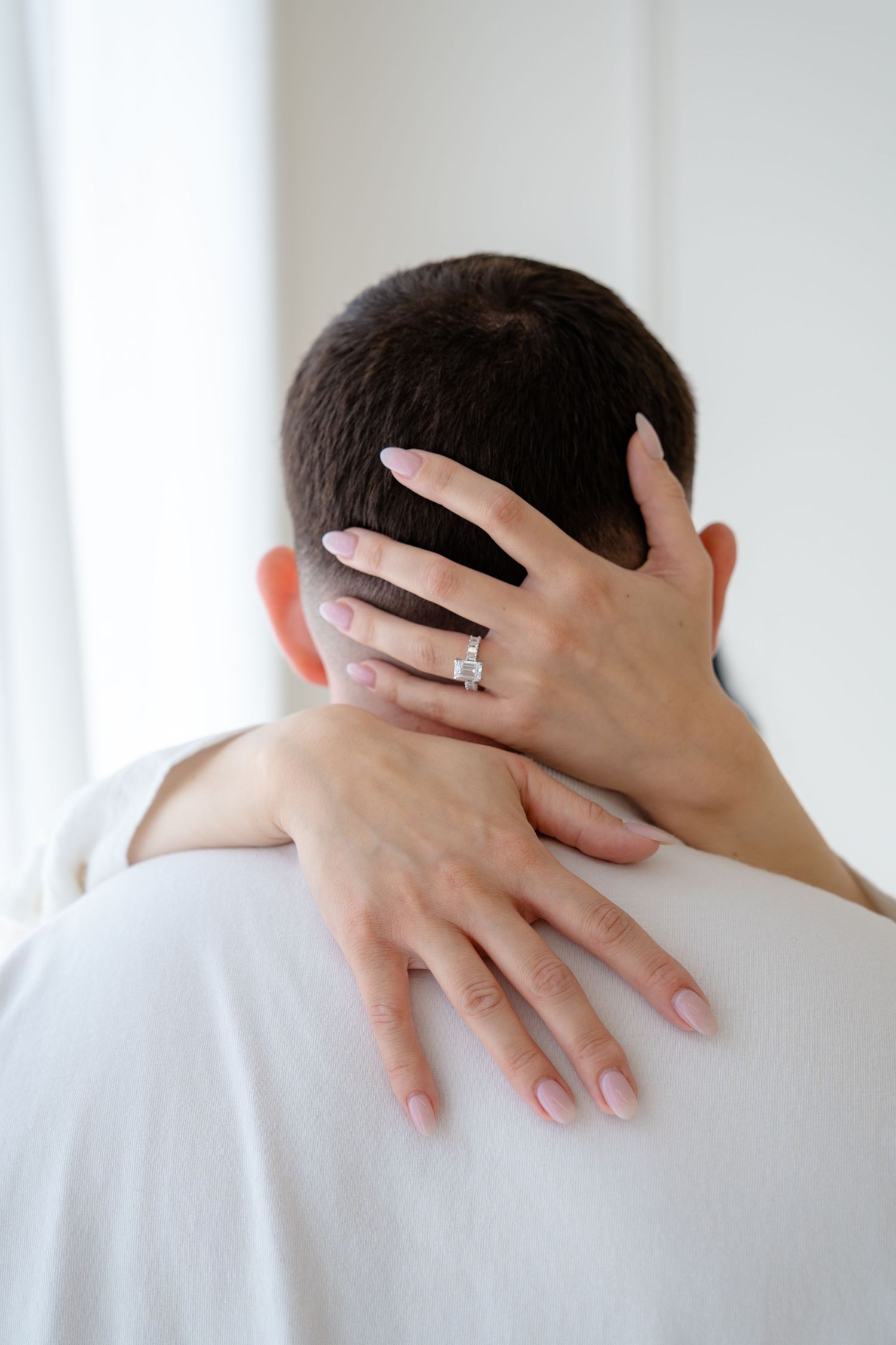 Woman wearing diamond engagement ring embracing man from behind, showing manicured pink nails against his neck.