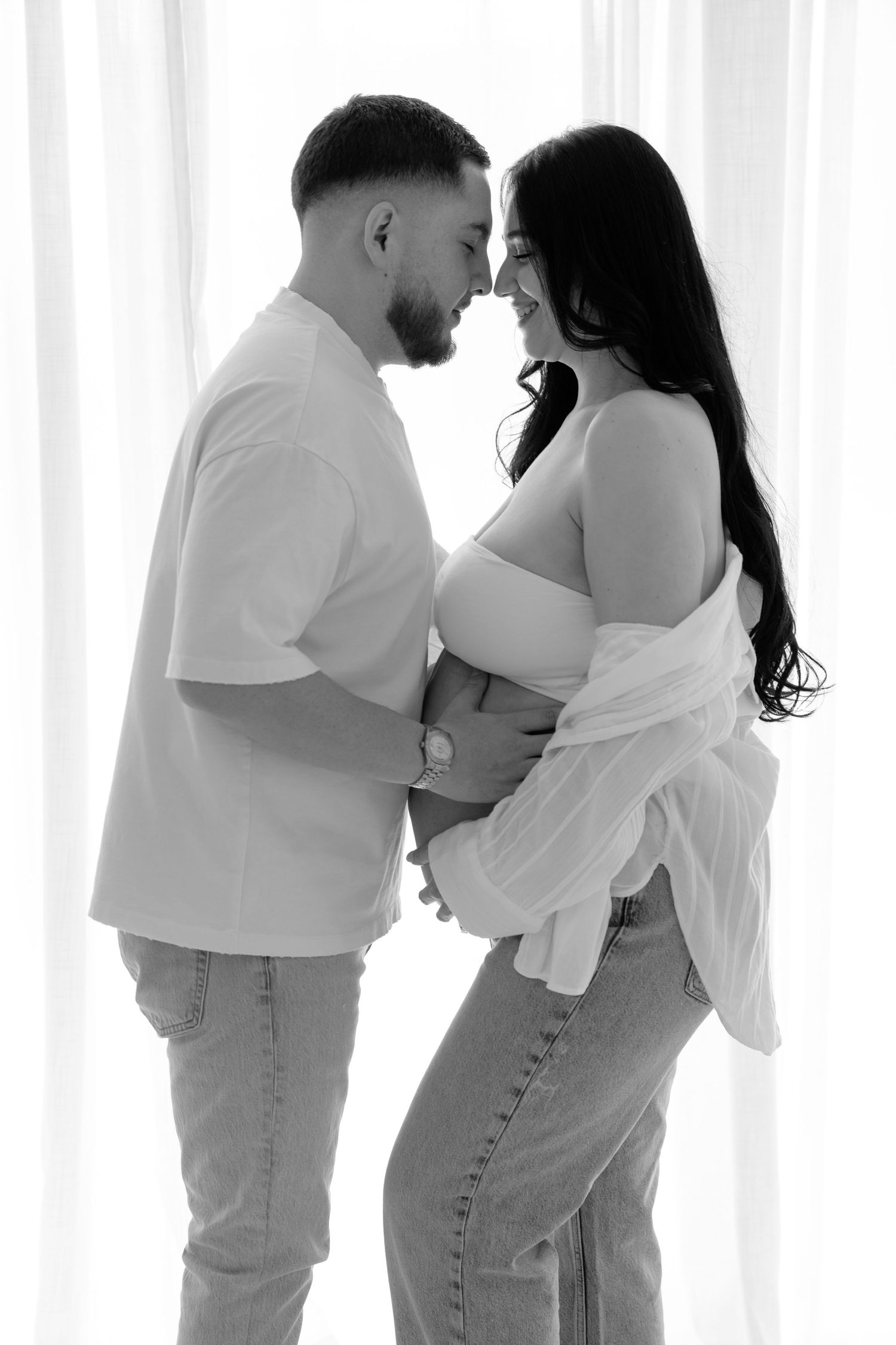 Black and white maternity photo of a couple tenderly touching foreheads while cradling a pregnant belly near sheer curtains.