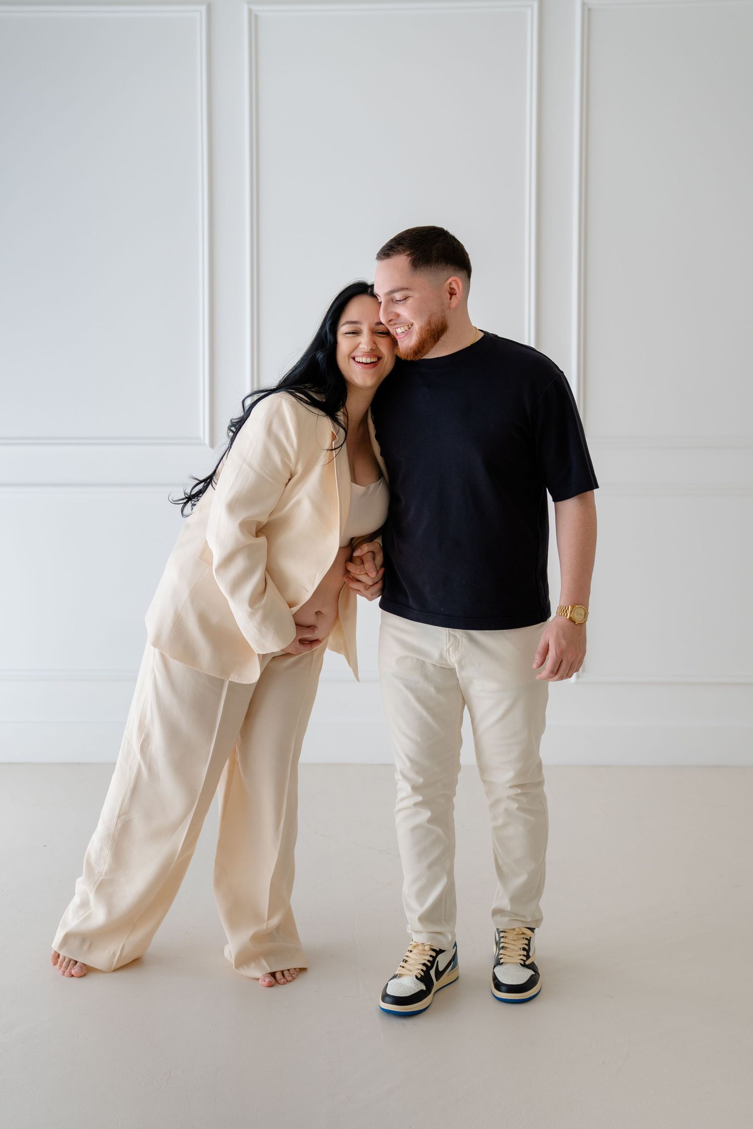 Happy couple posing together in a bright white studio, woman in cream suit leaning on man in black t-shirt and white pants.
