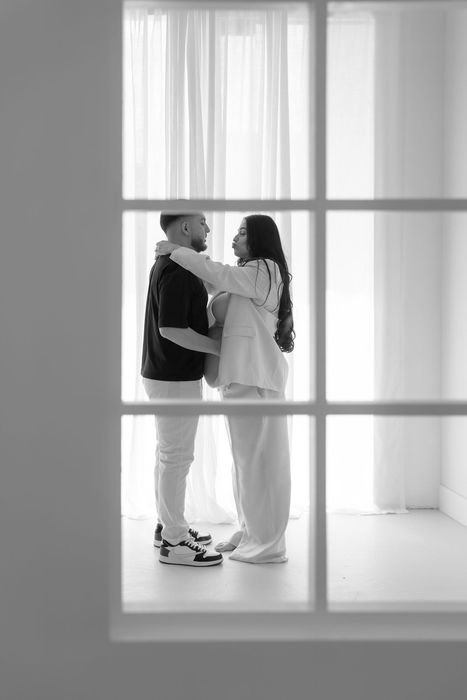 Black and white photo of a couple embracing and gazing at each other in front of sheer white curtains.