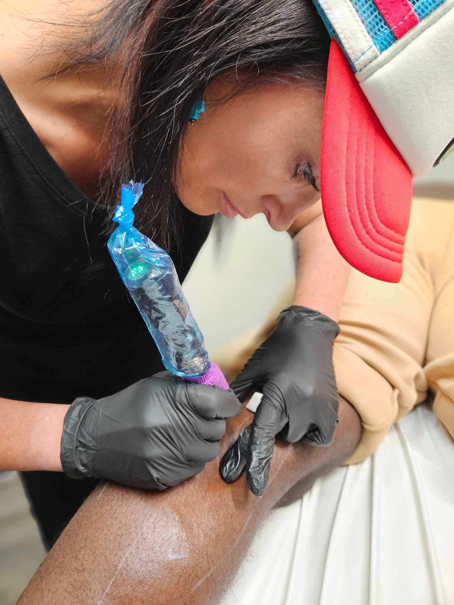 Tattoo artist wearing black gloves applies colorful ink with a wrapped tattoo machine on a client's arm.