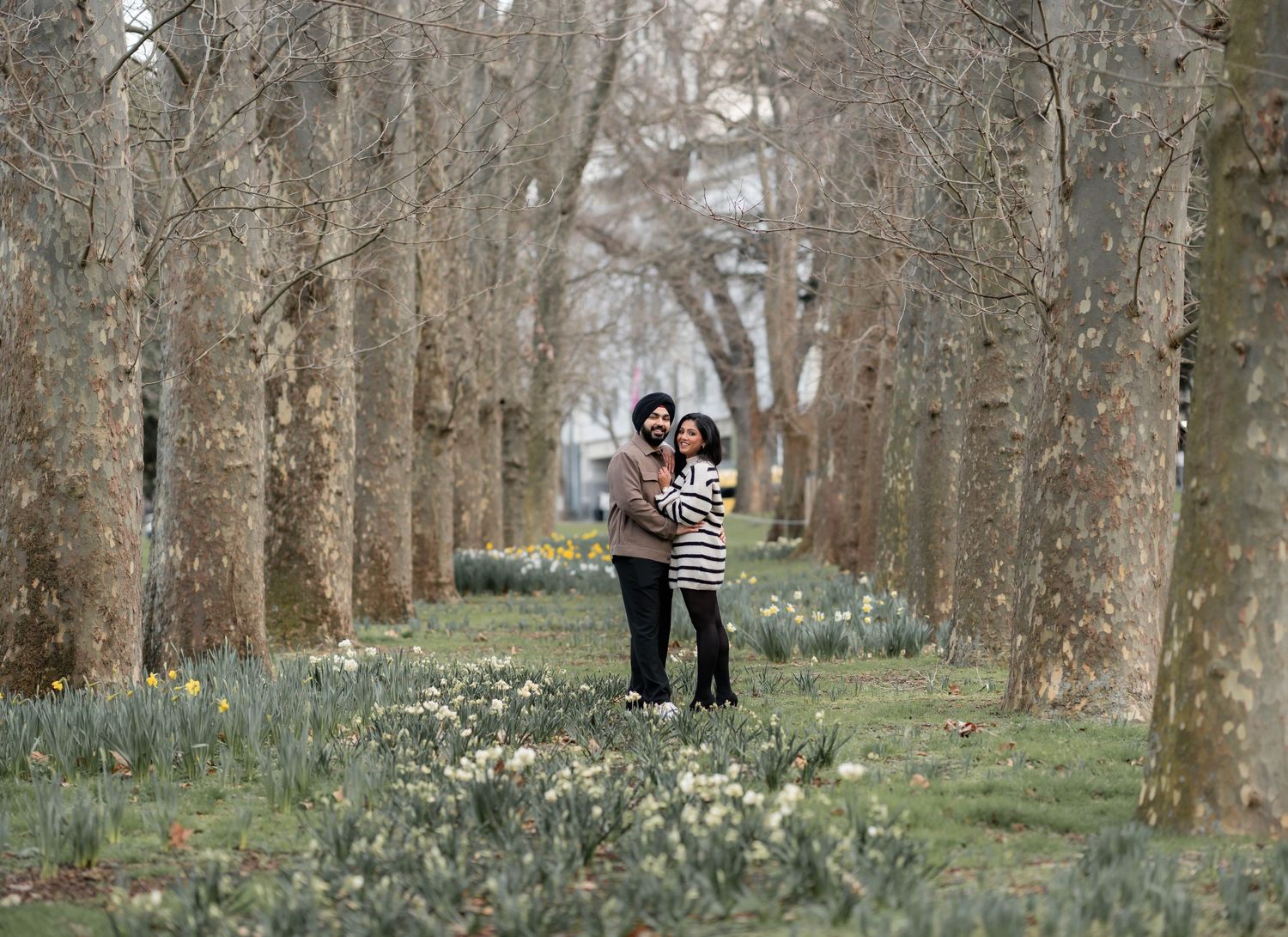 A couple embraces in a scenic tree-lined path with blooming spring flowers in the foreground.