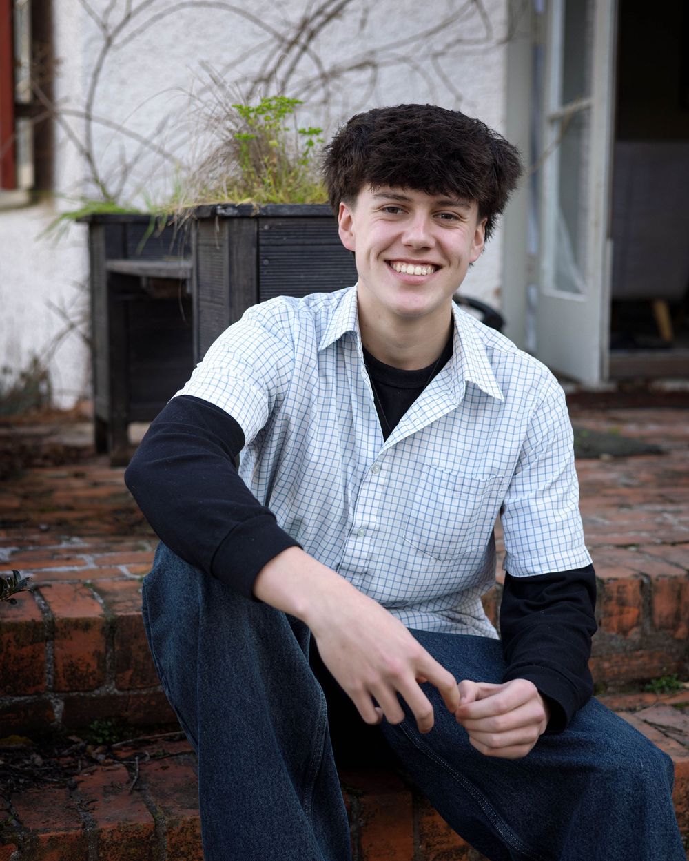 A young person wearing a checkered shirt and black undershirt sits on brick steps in front of a house with potted plants.