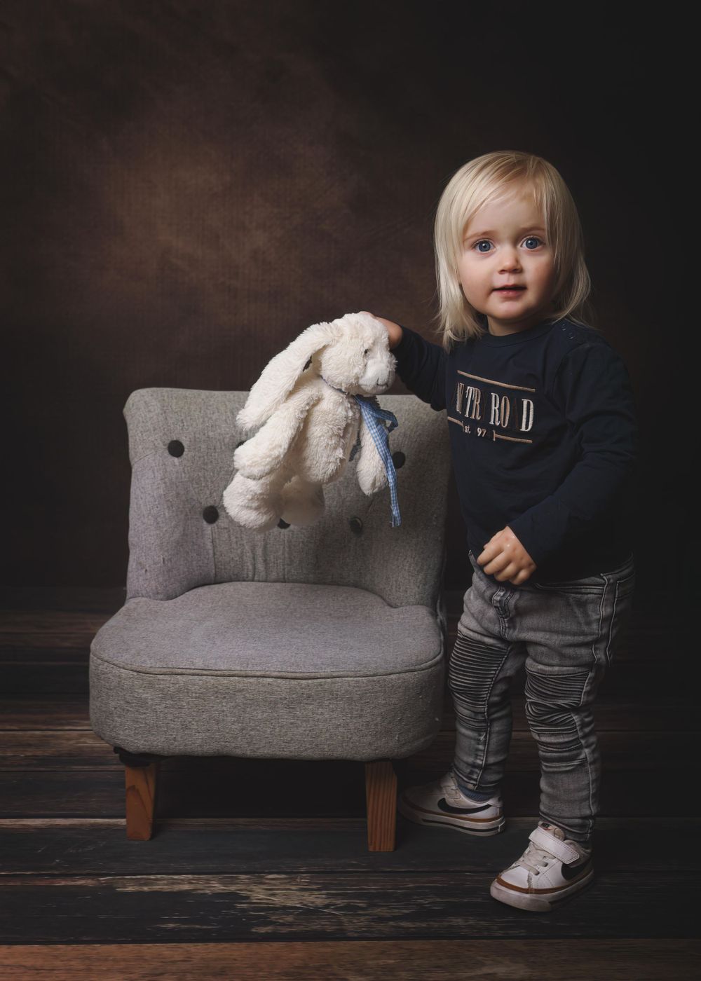Painterly portrait of young boy standing next to a small chair holding a bunny toy.