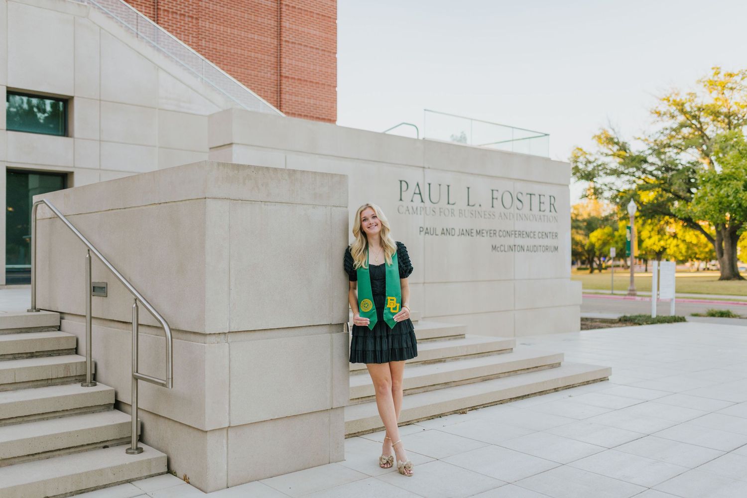 Blonde graduate in a black dress with a green Baylor stole standing outside the Paul L. Foster campus building.