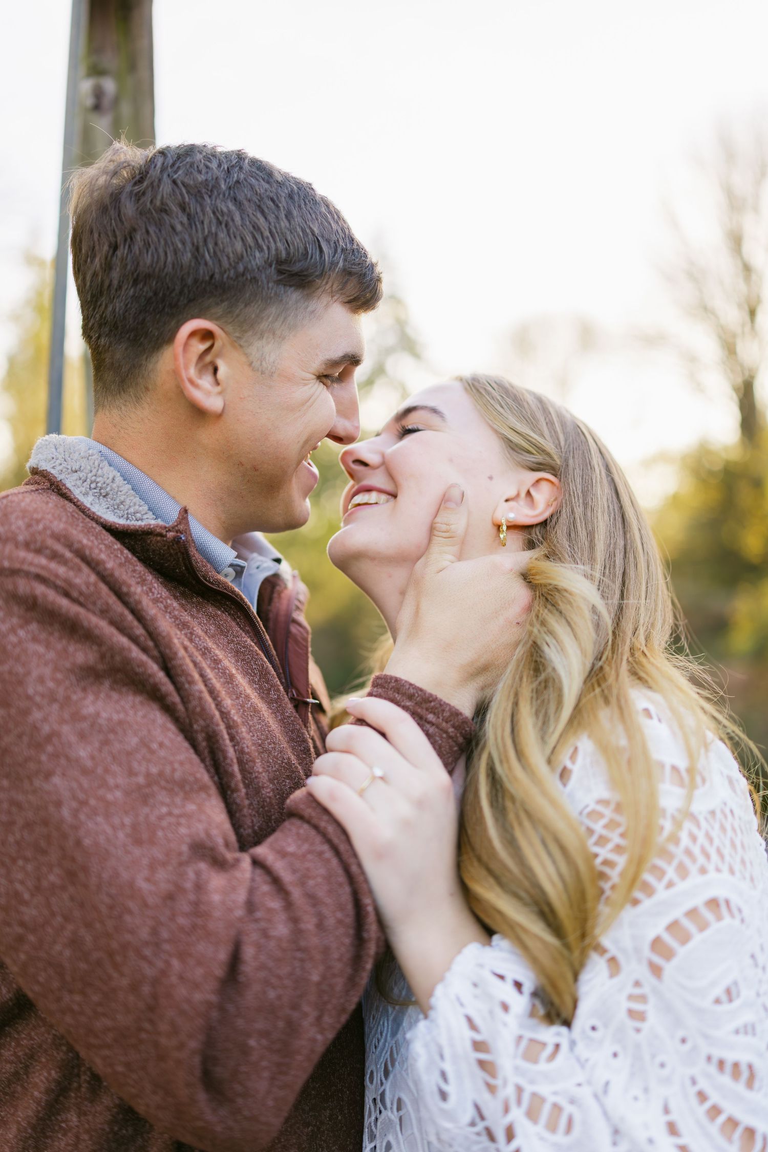 Couple laughing together outdoors, man in brown sweater gently holding womans face in warm light.