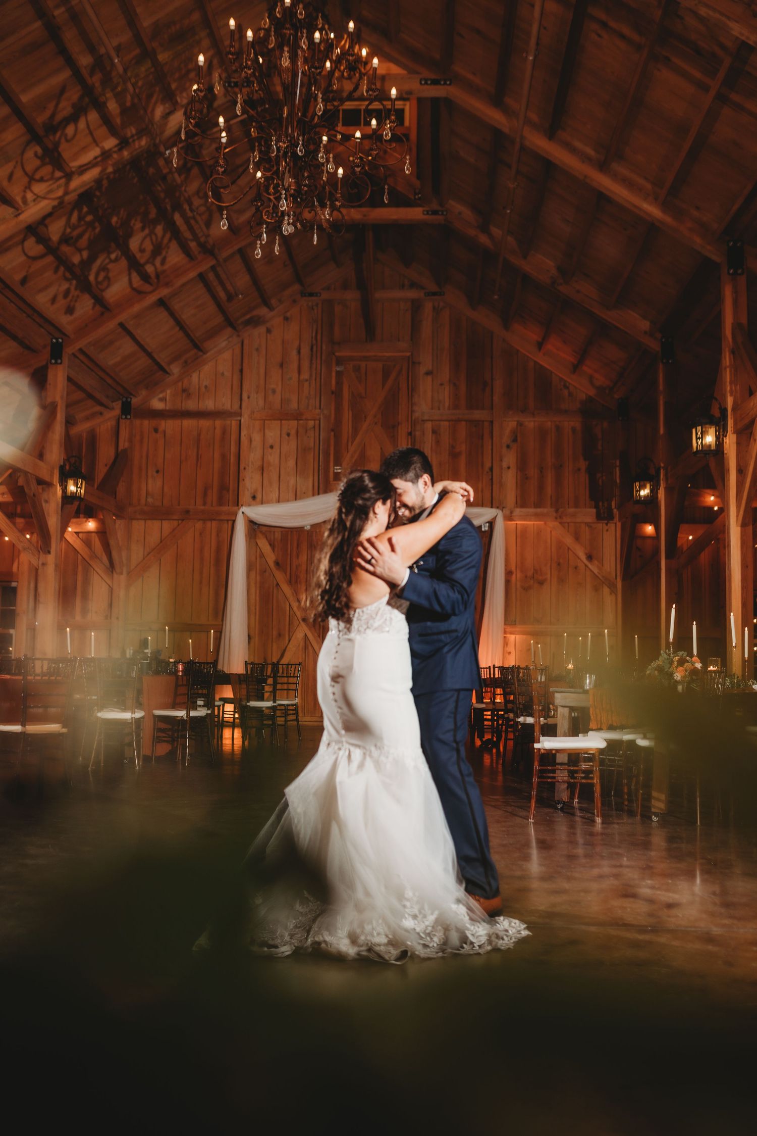 Romantic dance in a rustic barn venue under a grand chandelier with ambient lighting.