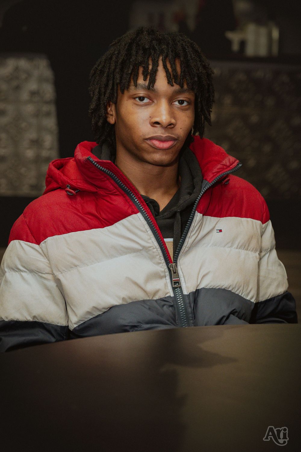Young man with dreadlocks wearing a red, white, and gray Tommy Hilfiger puffer jacket seated indoors.
