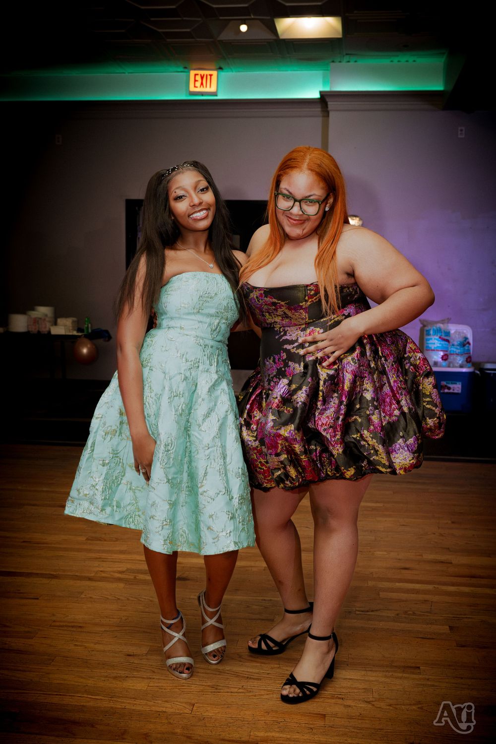 Two women in elegant dresses posing together at a formal event on a wooden dance floor.