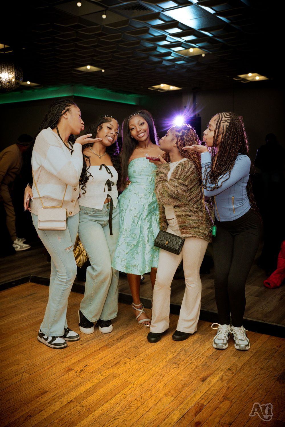 Five young women posing together and smiling at a party event on a wooden dance floor.